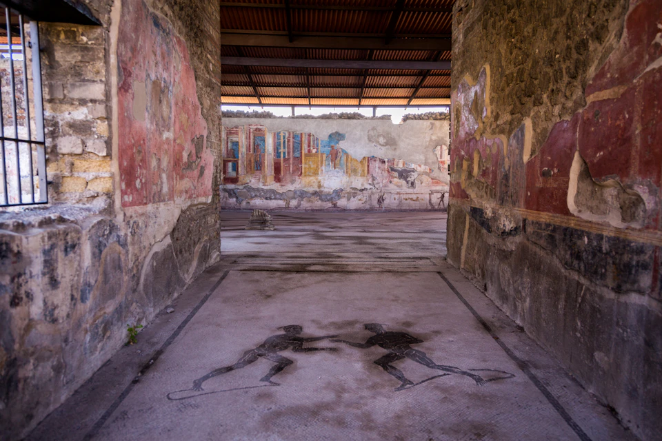 Ancient ruins with wall frescoes and a floor mosaic depicting two figures fighting; open-roof structure visible above.
