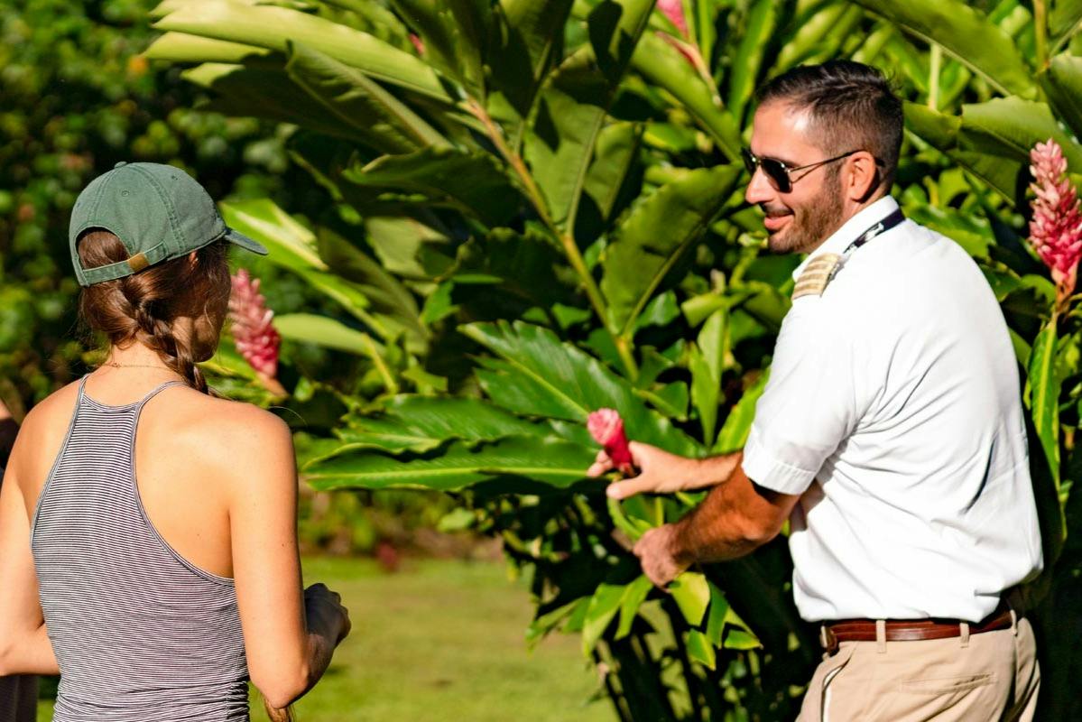 A man in a white shirt and sunglasses standing outdoors near large green leaves and pink flowers, smiling.