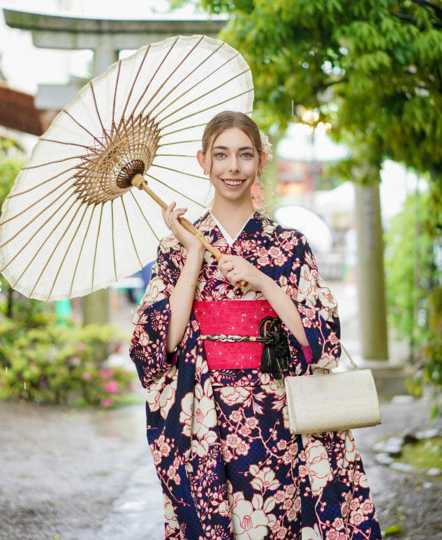 A woman wearing a floral kimono holds a parasol and a small purse, standing outdoors near greenery and a torii gate.