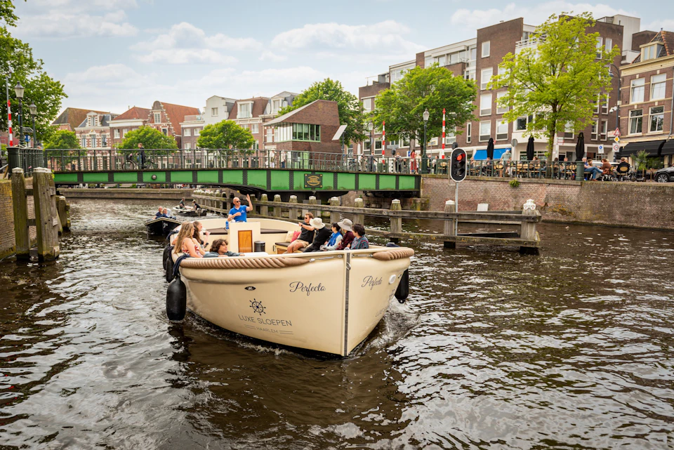 People enjoying a boat ride on a canal with a green bridge and residential buildings in the background.