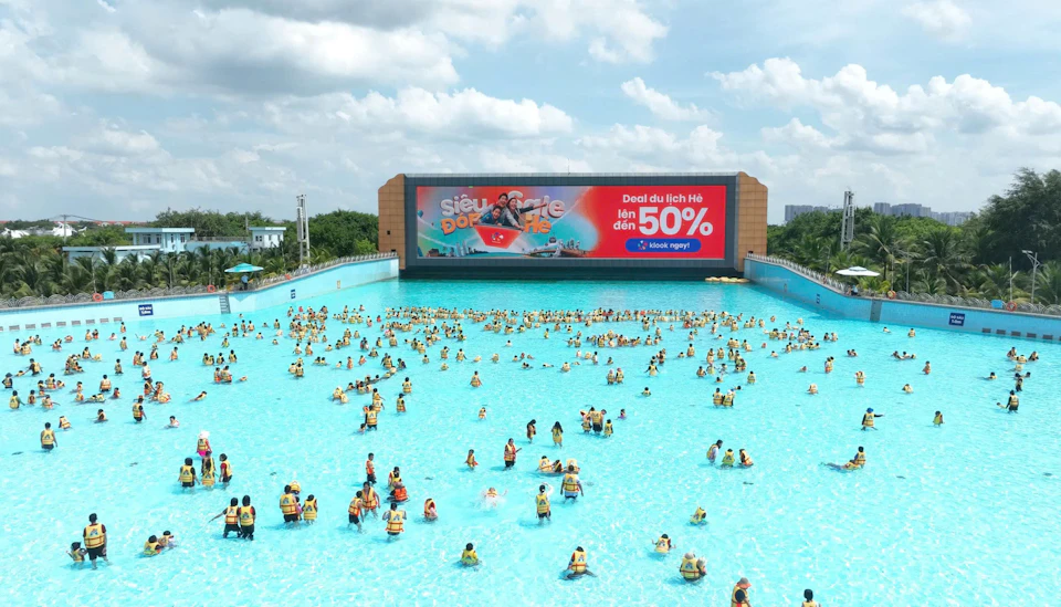 A large wave pool with many people wearing life vests. A billboard with an advertisement is visible in the background.
