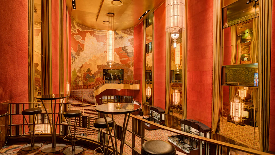 Luxurious art-deco interior with red walls, intricate murals, grand chandeliers, tiered seating, and bar tables with high stools.
