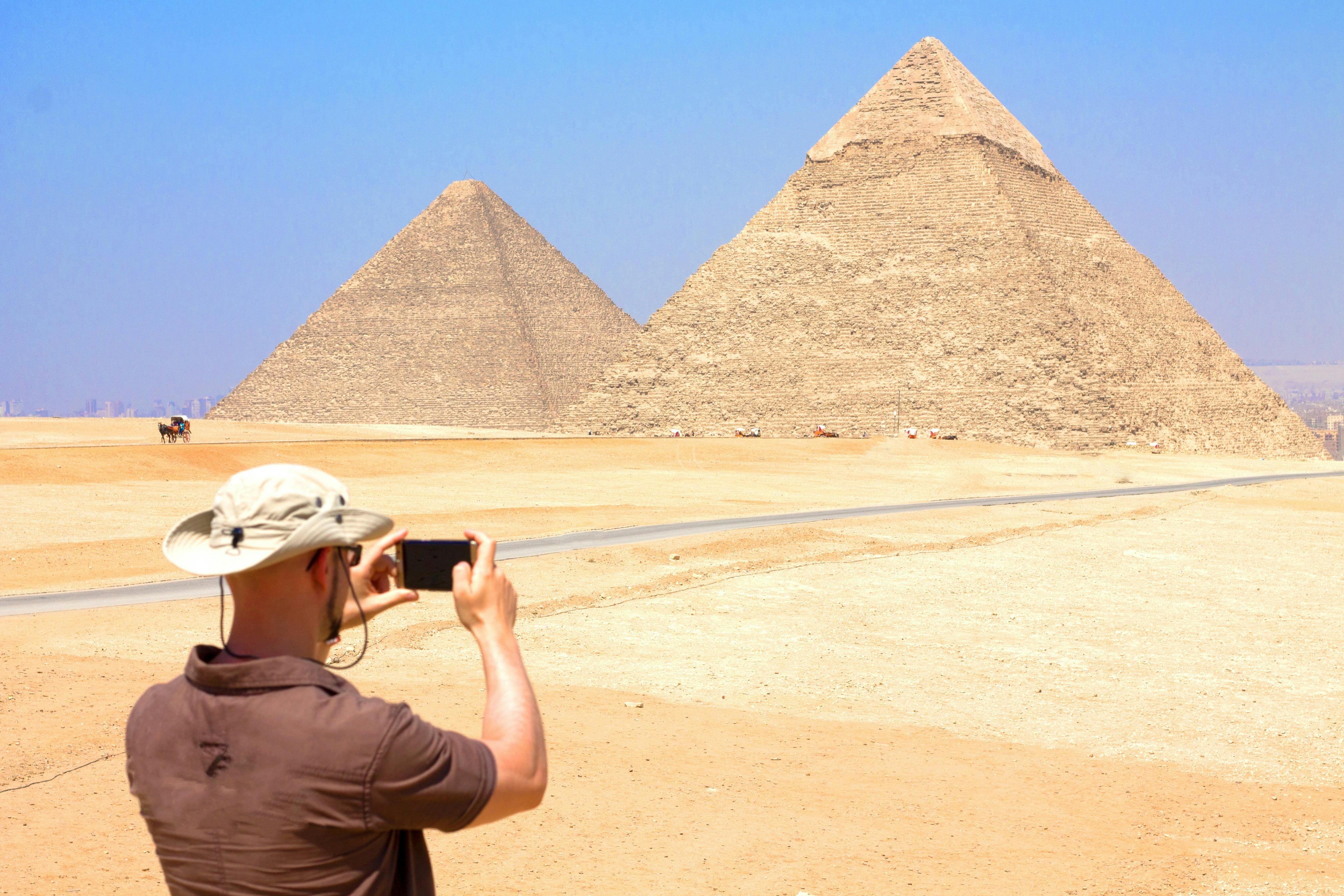A person wearing a hat takes a photo of the Pyramids of Giza against a clear blue sky.