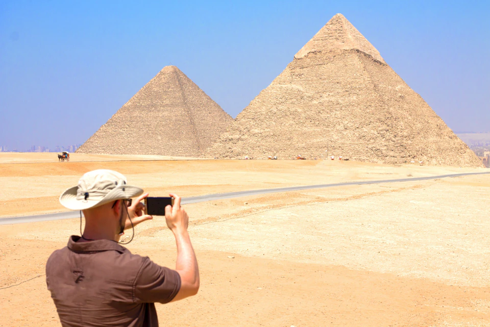 A person wearing a hat takes a photo of the Pyramids of Giza against a clear blue sky.