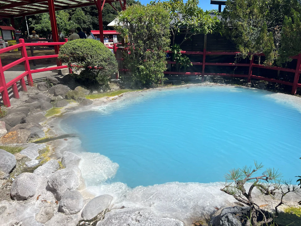 A serene hot spring with blue water surrounded by rocks, plants, and a red wooden fence under clear skies.
