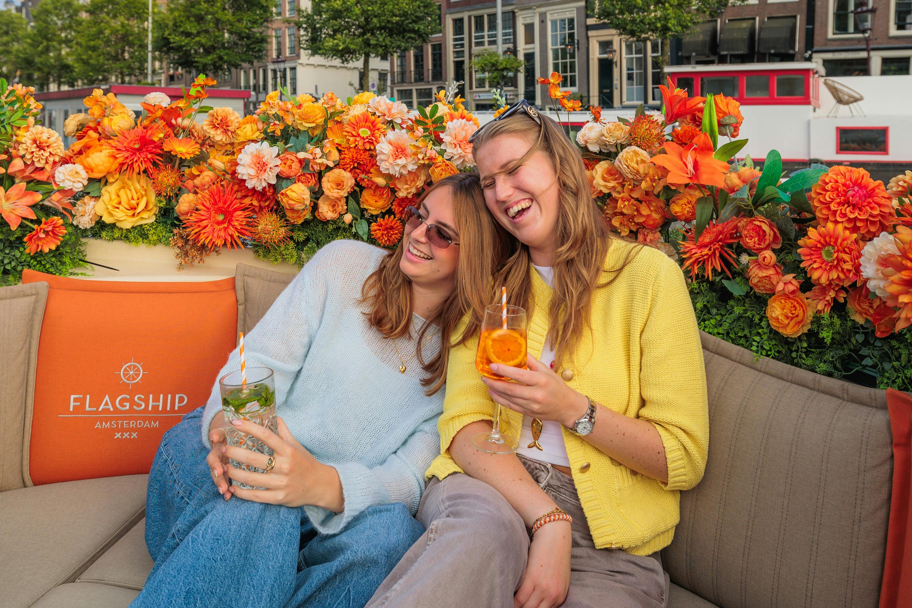 Two women smiling and sitting together outdoors, holding drinks, with colorful orange flowers and a street scene in the background.