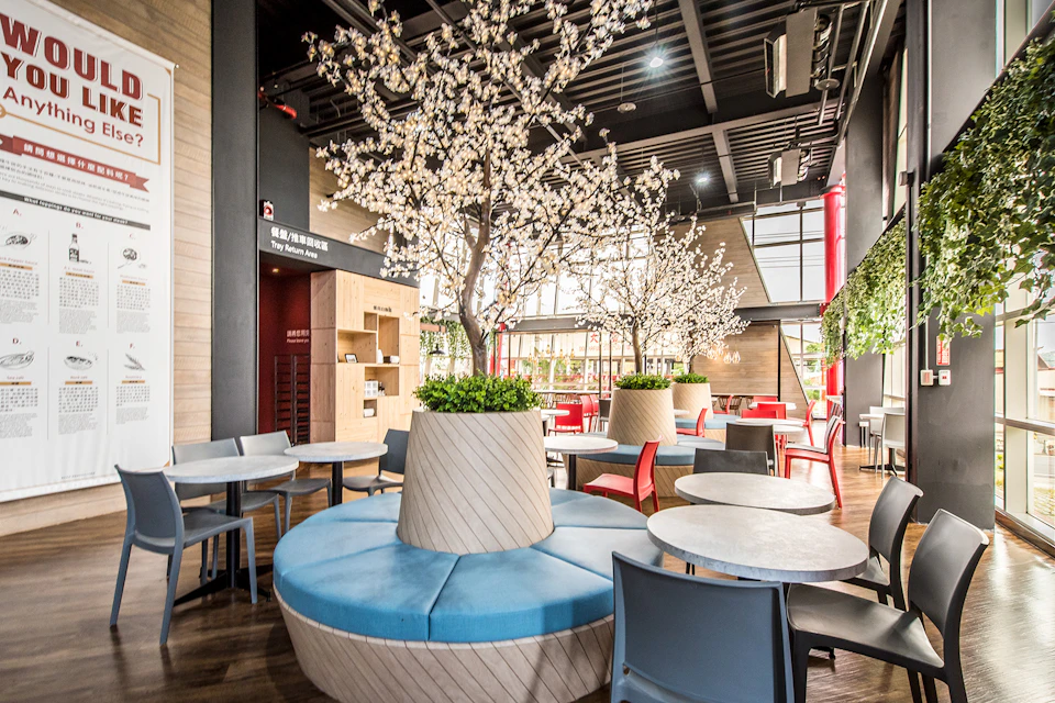 A modern café with blue cushioned seating, white blossom trees as decor, and tables surrounded by chairs under a high ceiling.