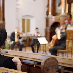 En person med hästsvans ser en stråkkvartett uppträda i en kyrka från en träbänk.