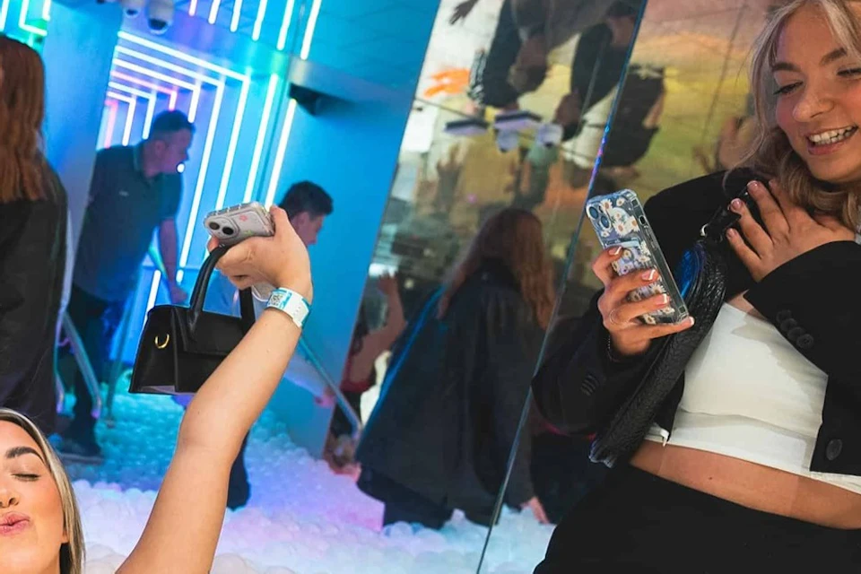 Two women enjoying a ball pit filled with white balls in a vibrant, neon-lit indoor setting.