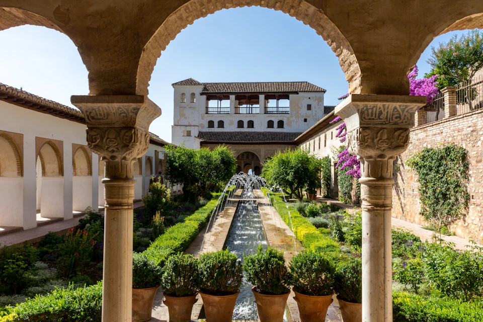 Ornate stone arches frame a courtyard garden with a central fountain, lush greenery, and a large building in the background.
