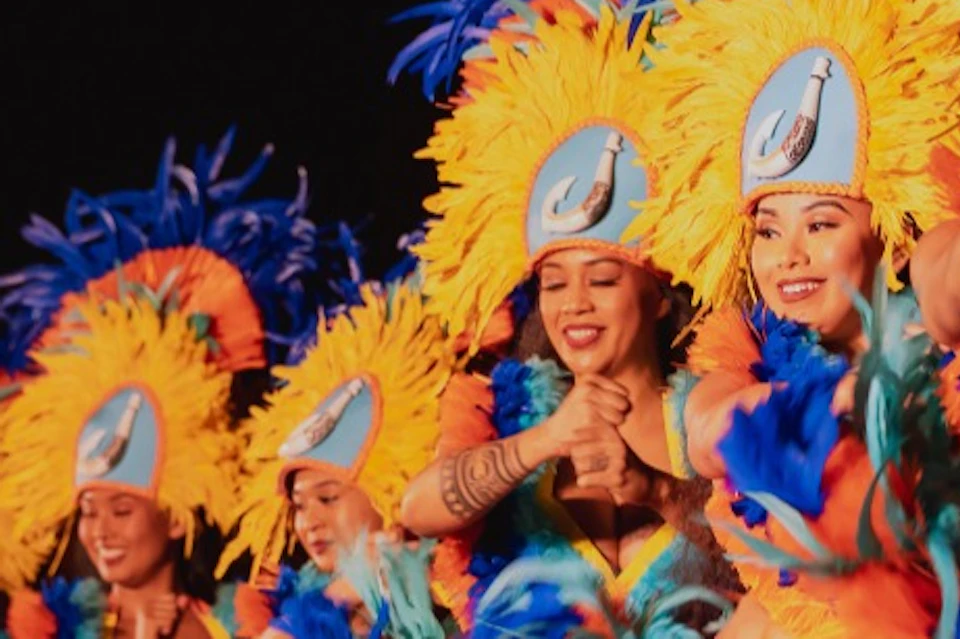 A group of performers in brightly colored costumes with feathered headdresses dancing in unison on stage.