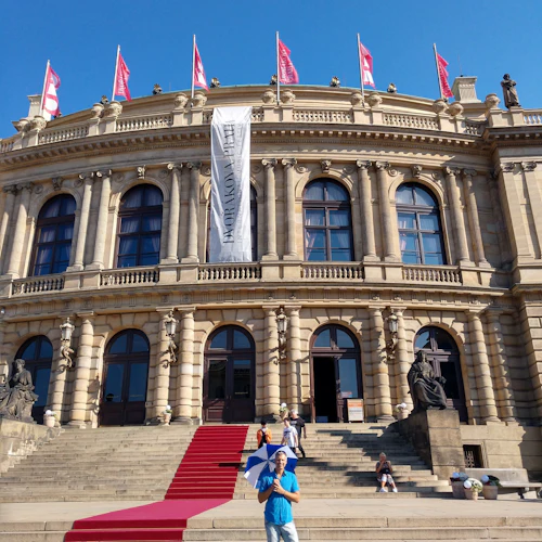En person med ett paraply står framför en storslagen byggnad med en röd matta, flera flaggor och flera statyer.