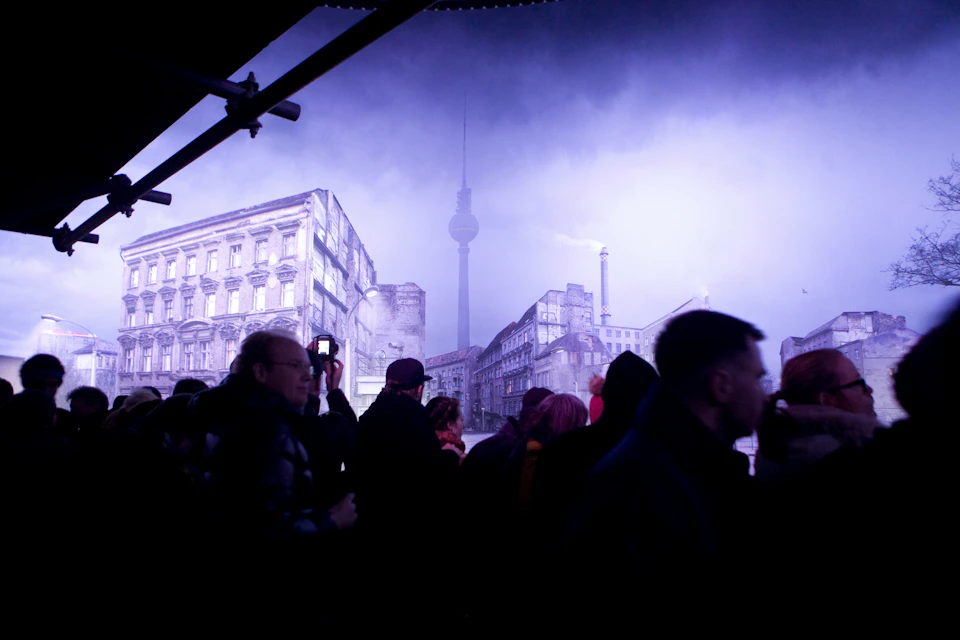 Silhouettes of people stand in front of illuminated buildings, with a tall tower visible in the background under a cloudy sky.