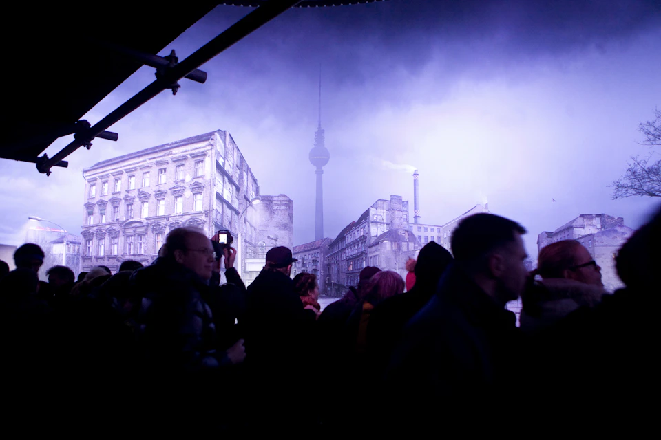 Silhouettes of people stand in front of illuminated buildings, with a tall tower visible in the background under a cloudy sky.