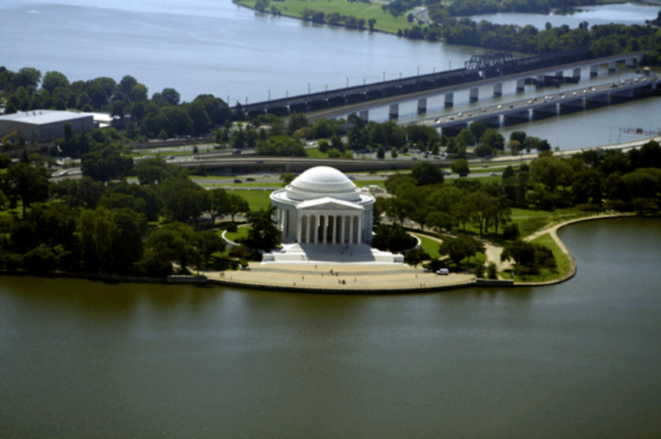 Aerial view of the Thomas Jefferson Memorial surrounded by water and greenery with a bridge and a highway in the background.