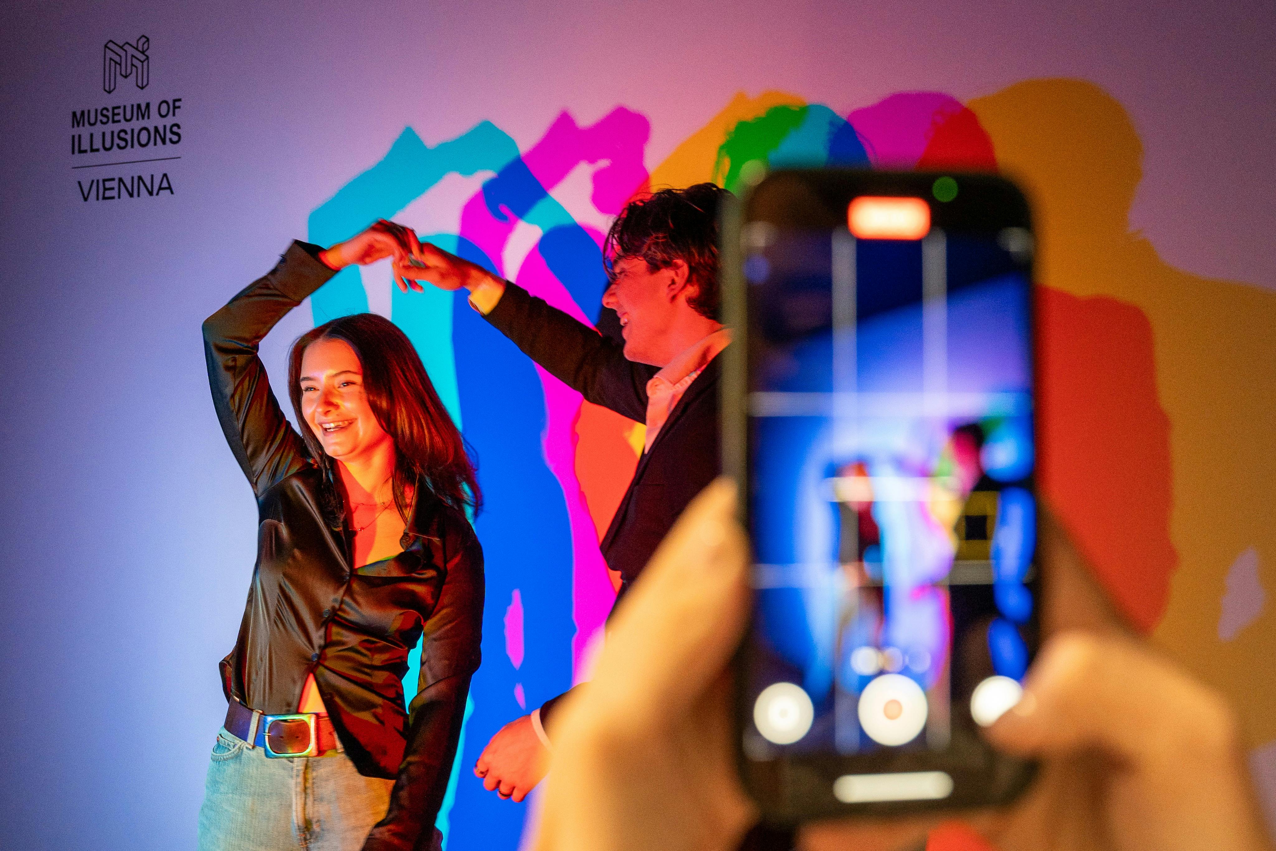 Duas pessoas dançam sob uma iluminação colorida no Museu das Ilusões em Viena, com um telemóvel a captar o momento.
