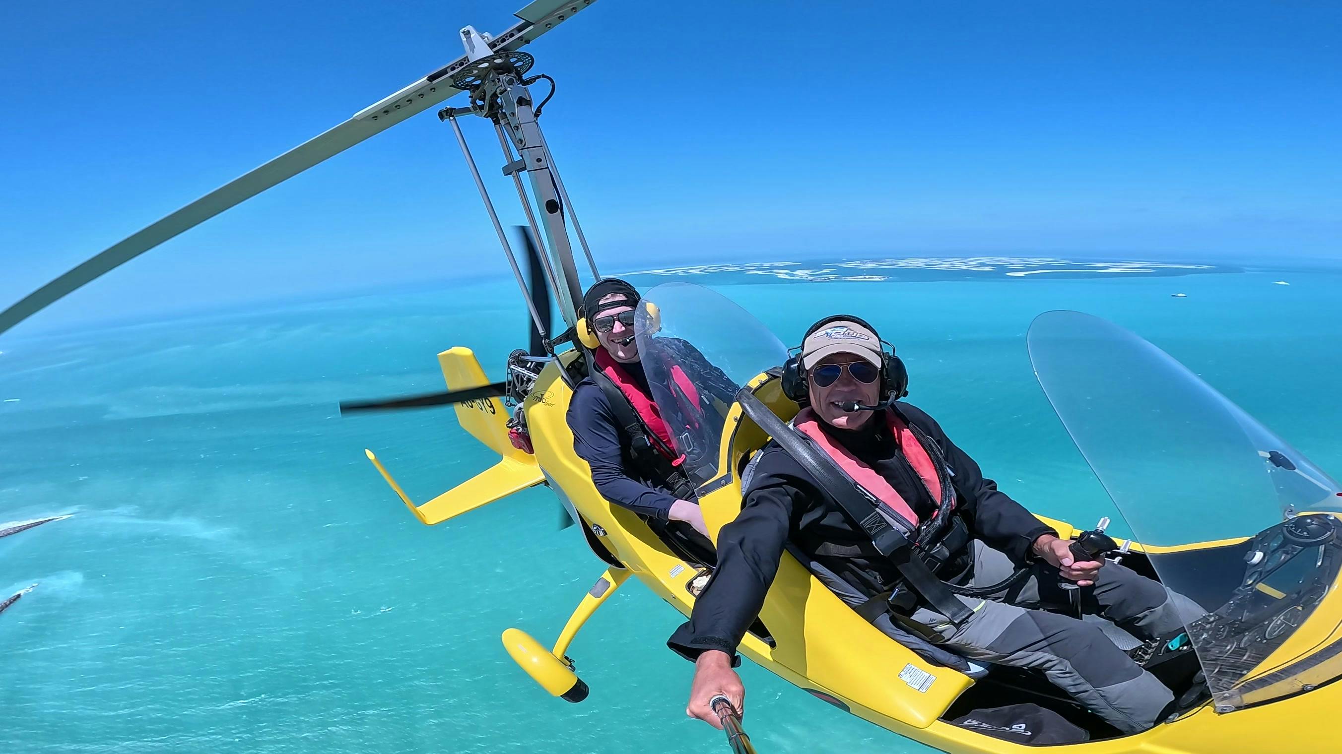 Duas pessoas num girocóptero amarelo a sobrevoar um oceano azul-turquesa sob um céu azul claro.