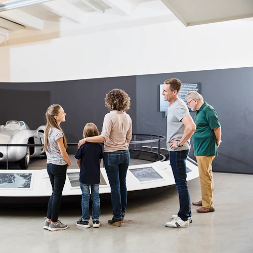 Fem personer beundrar en vintage racerbil i silver som visas på ett museum med informationsplaketter framför.