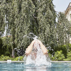 Person som kommer ut från en pool, stänker vatten, med frodiga träd och en byggnad i bakgrunden.