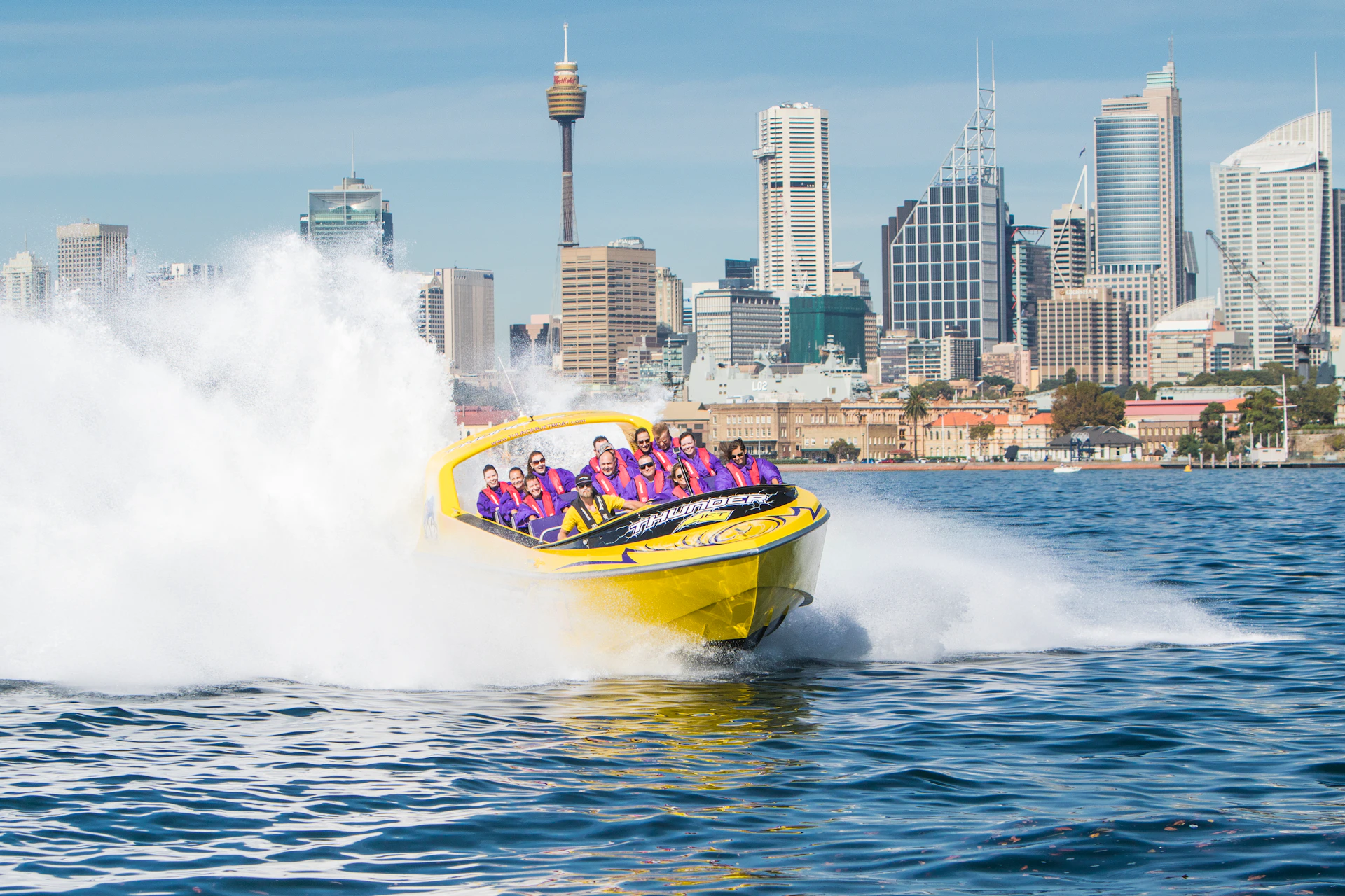 A yellow speedboat with passengers creates large splashes on the water, with a city skyline, including a tower, in the background.