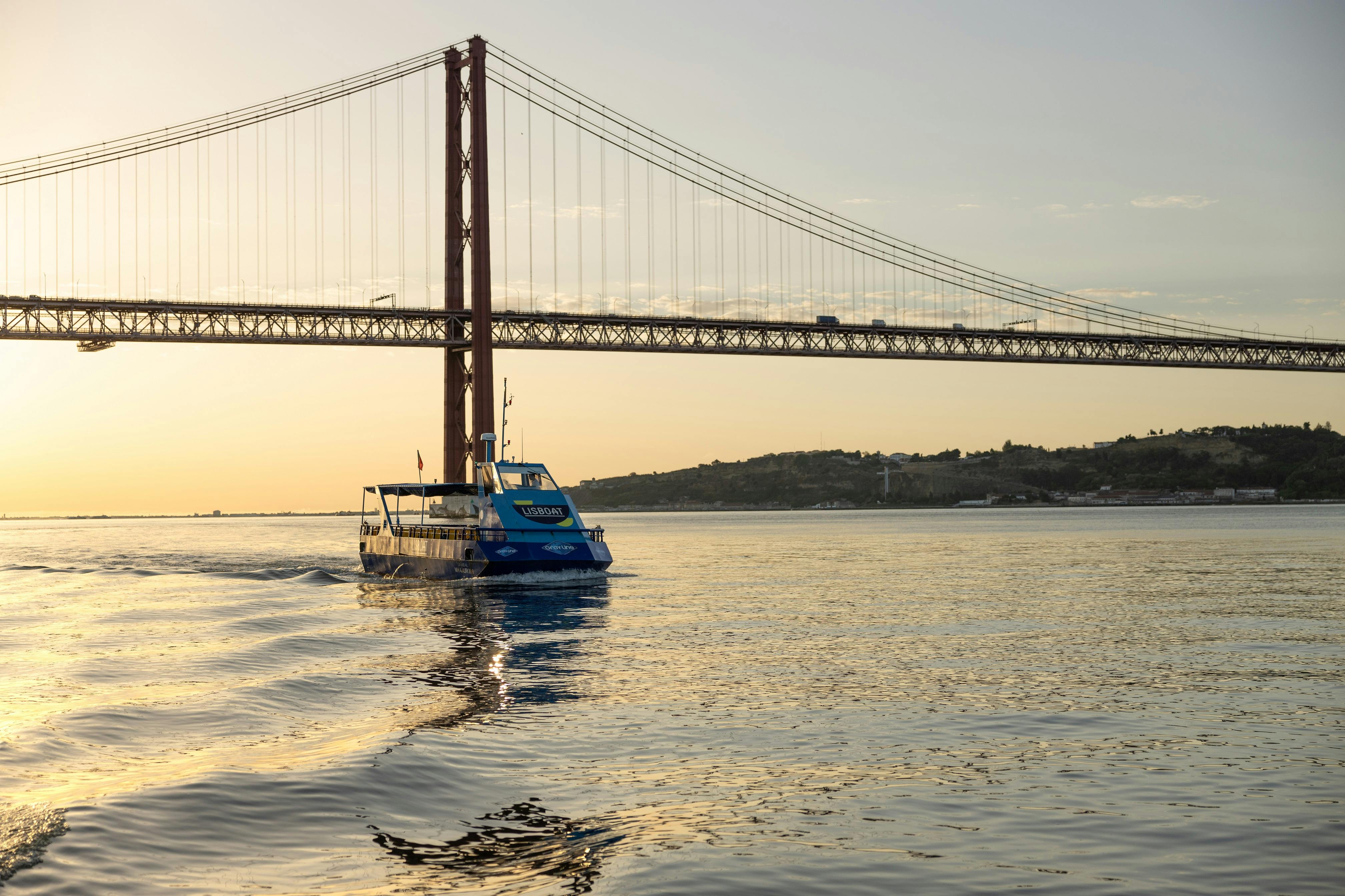 Eine blaue Bootstour auf ruhigem Wasser mit einer großen Hängebrücke und Hügeln im Hintergrund bei Sonnenuntergang.