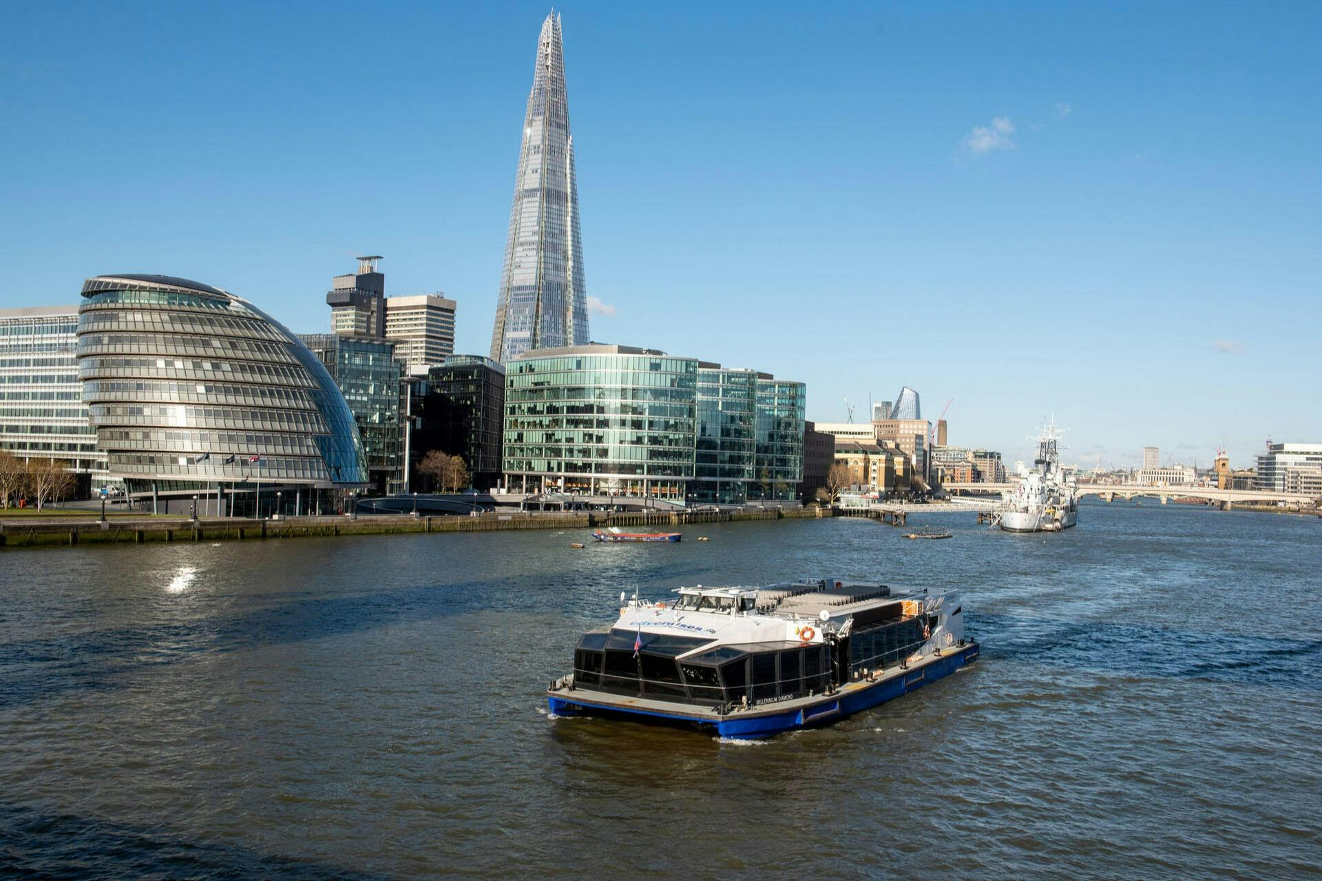 A boat navigates the River Thames with modern buildings, including The Shard, visible against a blue sky.