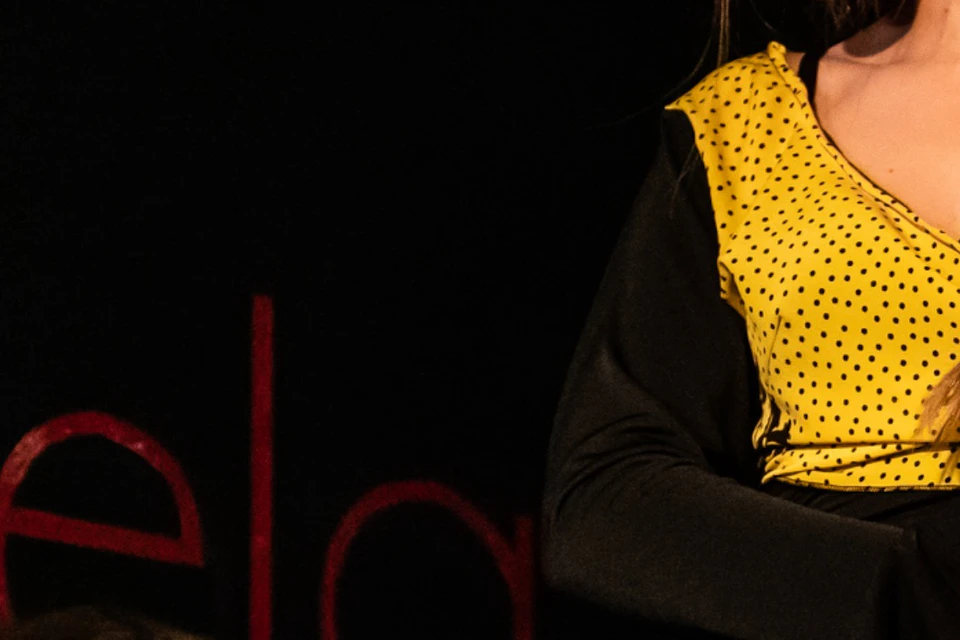 A performer in a yellow and black dress stands on stage, while another person claps in the background under dim lighting.