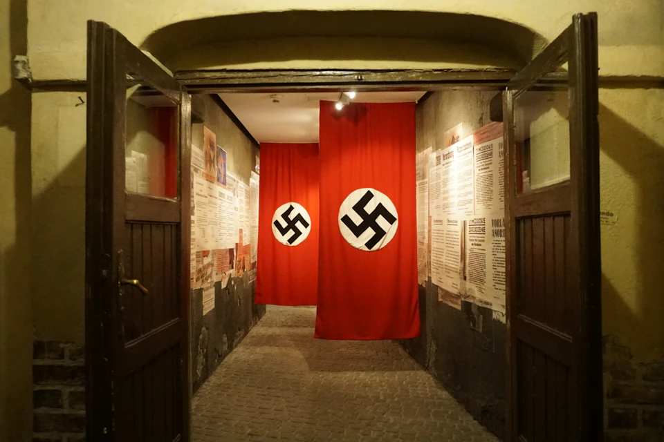 A narrow hallway displays two large red banners with black swastikas. The walls are covered with historical information and posters.