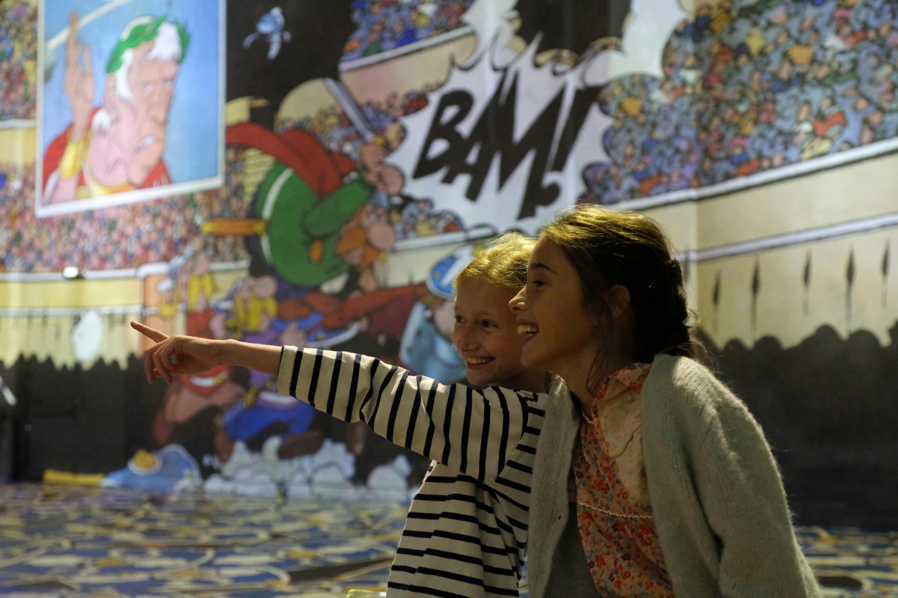 Dos nens somrient i assenyalant davant d'un colorit mural d'estil còmic amb la paraula "BAM!" en un lloc destacat.