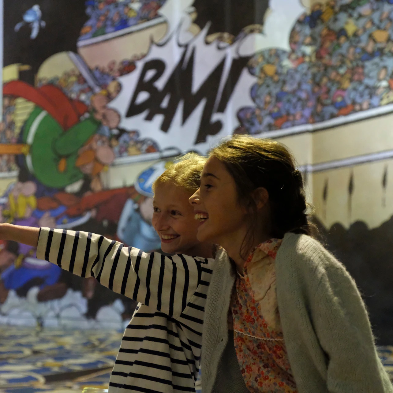 Two children smiling and pointing in front of a colorful comic-style mural with the word "BAM!" prominently displayed.