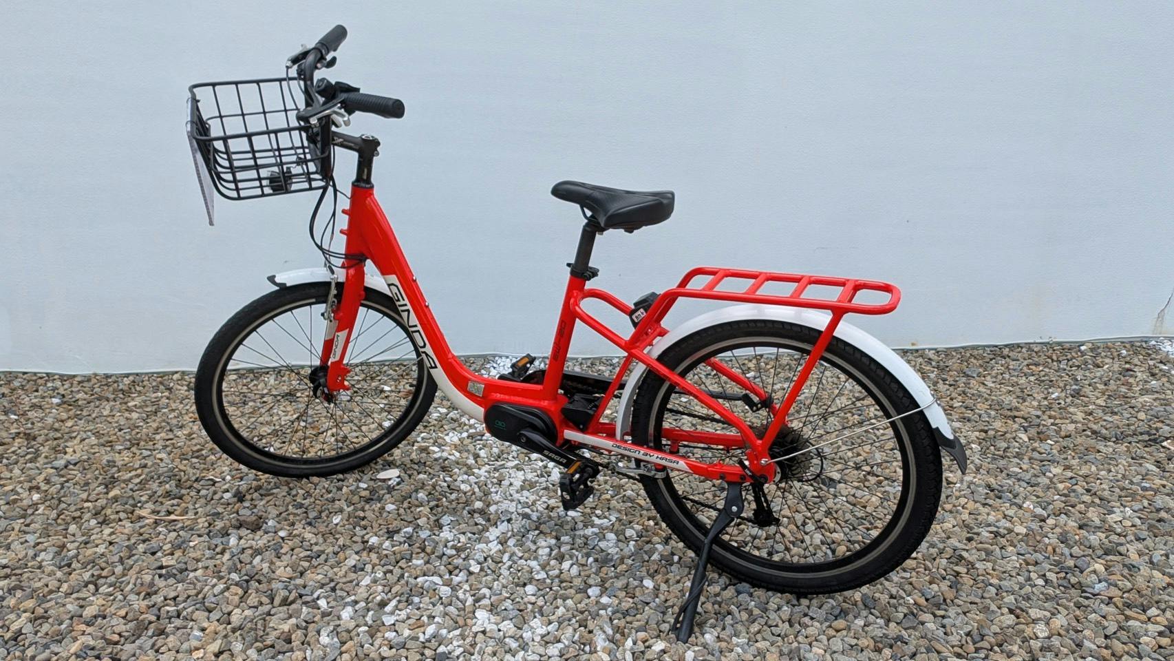 A red bicycle with a basket on the front and a rear cargo rack, parked on a gravel surface against a plain wall.