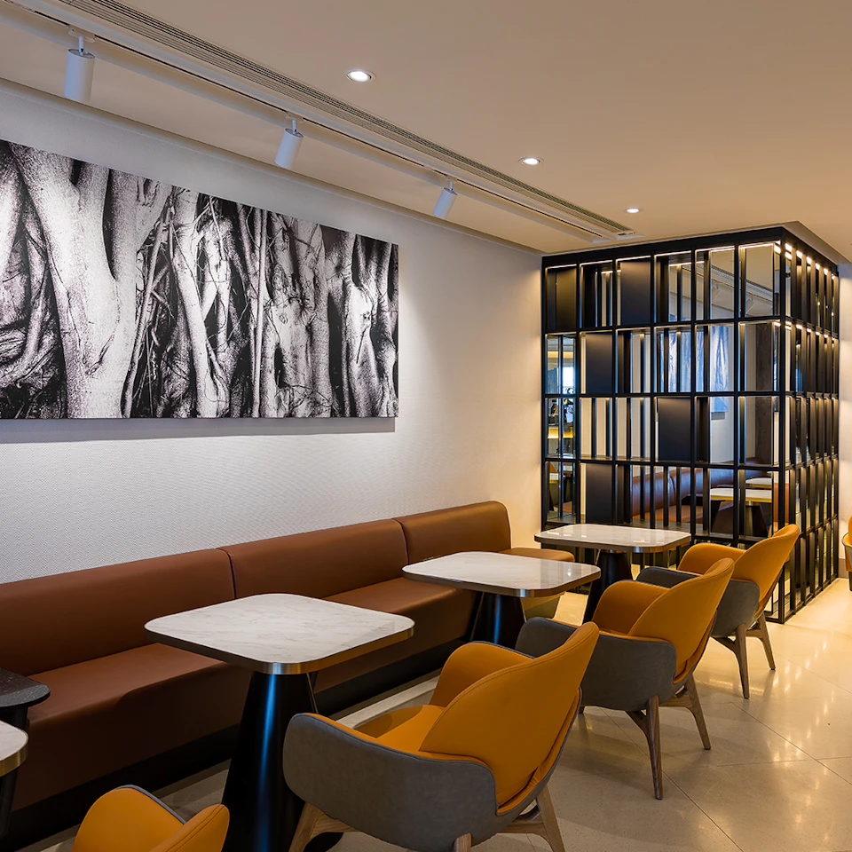 Modern café interior with mustard-yellow and grey chairs, marble-topped tables, a brown upholstered bench, and a black-and-white wall art.
