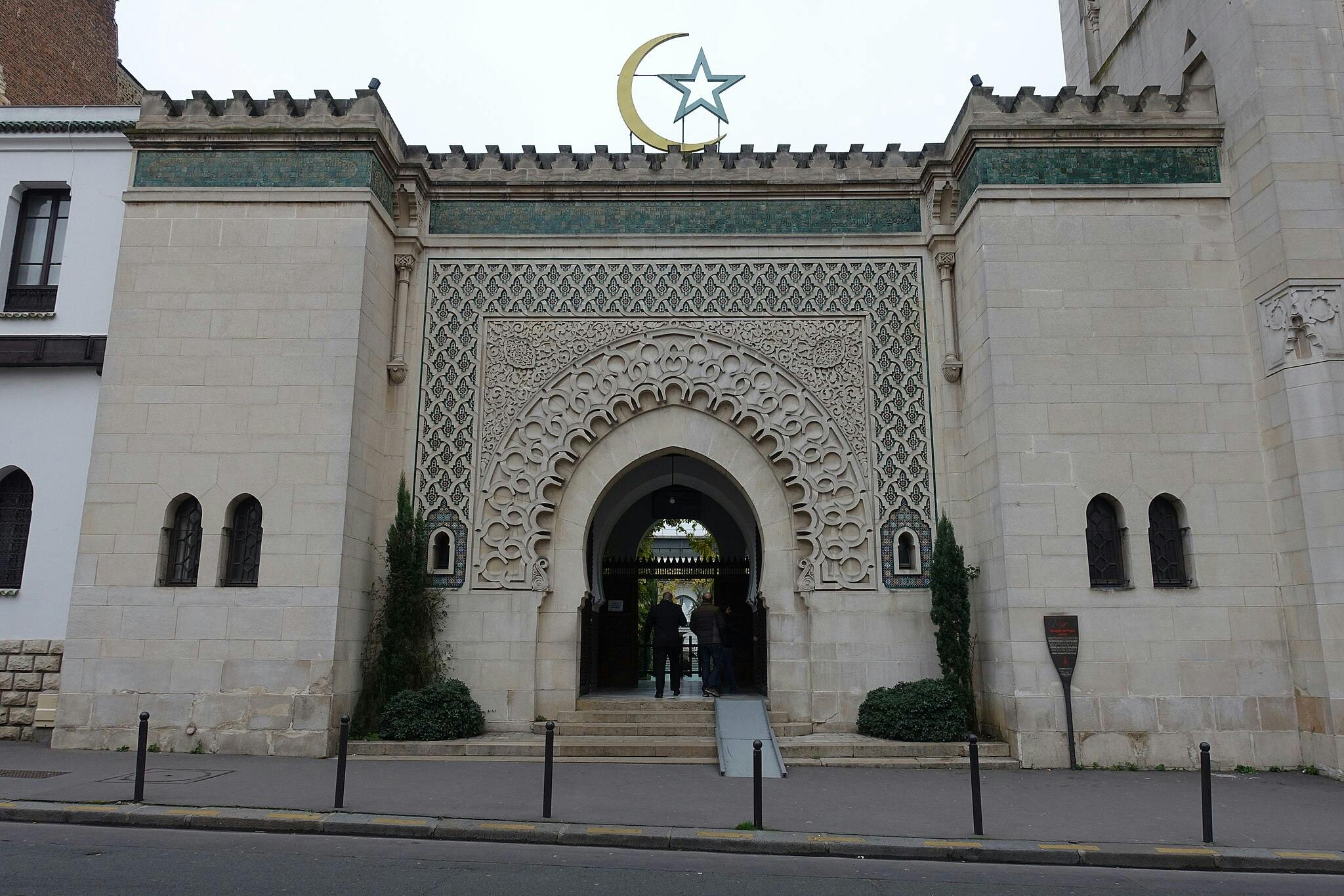 Ornate arched entrance of a mosque with a crescent and star on top, two figures entering, flanked by narrow windows and plants.