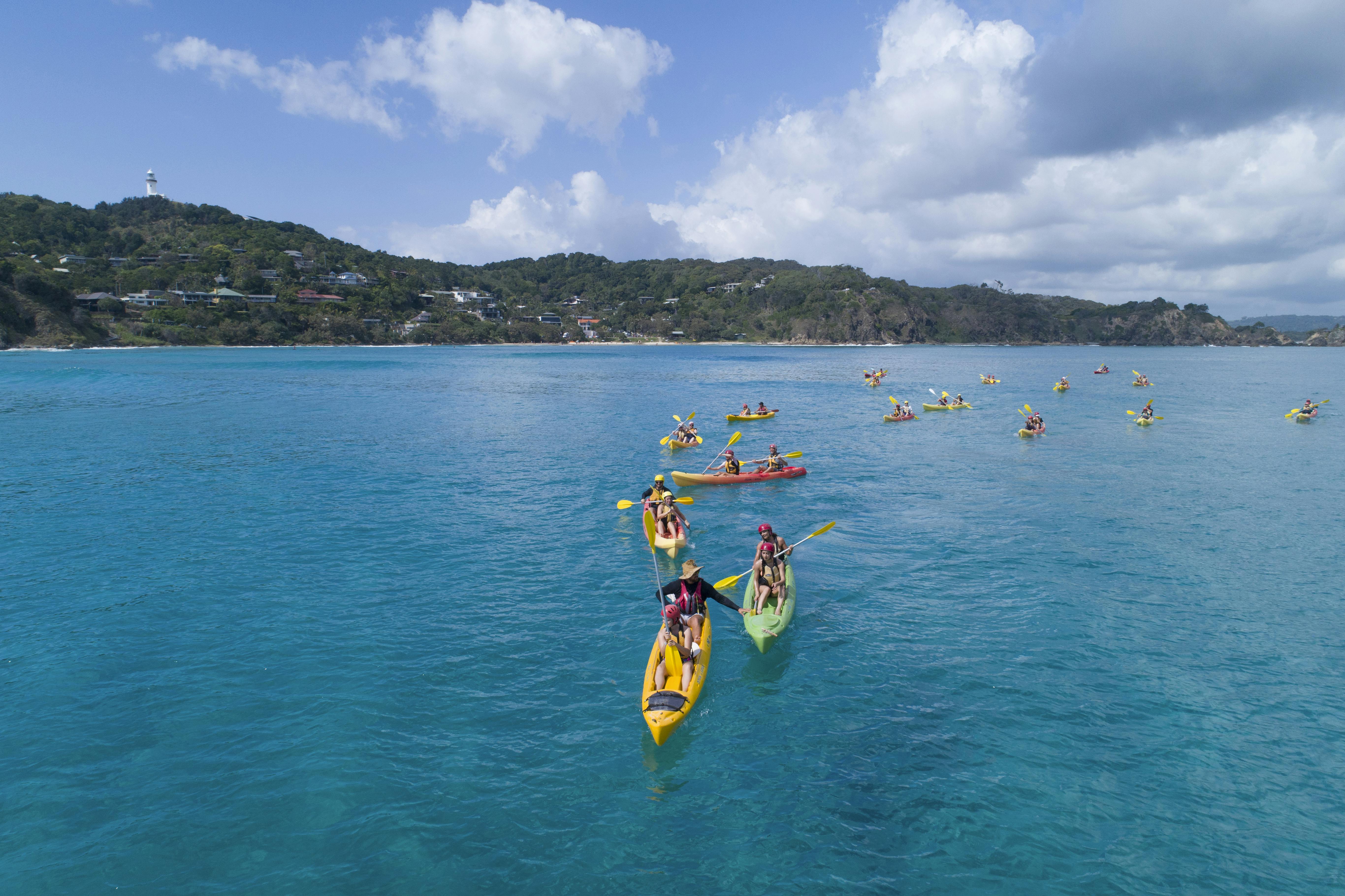 Groups of people kayaking on a calm, blue sea with a hilly coastline in the background under a partly cloudy sky.