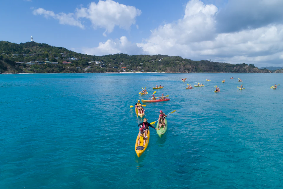Groups of people kayaking on a calm, blue sea with a hilly coastline in the background under a partly cloudy sky.