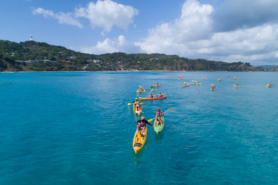 Groups of people kayaking on a calm, blue sea with a hilly coastline in the background under a partly cloudy sky.