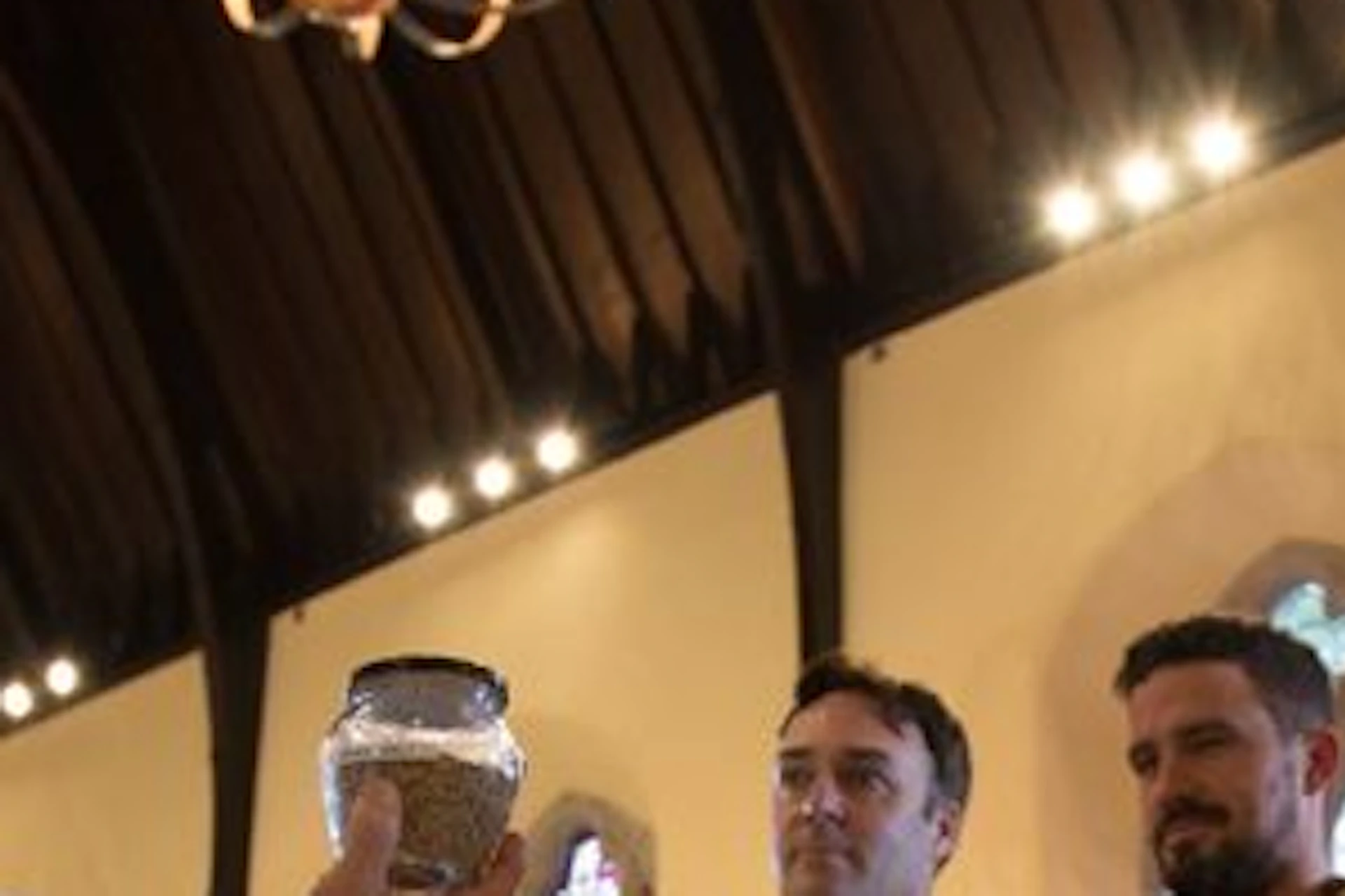A man in a teal vest explains something to a group of five people inside a building with wooden beams and barrels.