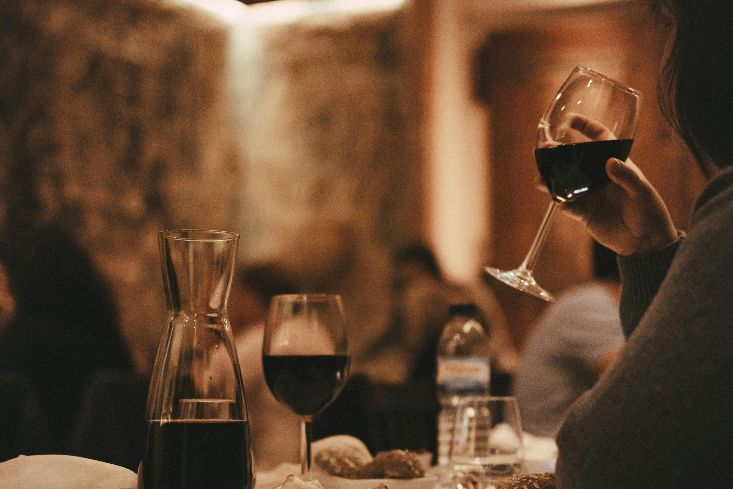 Ποτήρια κρασιού, μια κανάτα κόκκινο κρασί και ένα μπουκάλι νερό σε μια τραπεζαρία. Ένα χέρι που κρατάει ένα ποτήρι κρασιού στο παρασκήνιο.