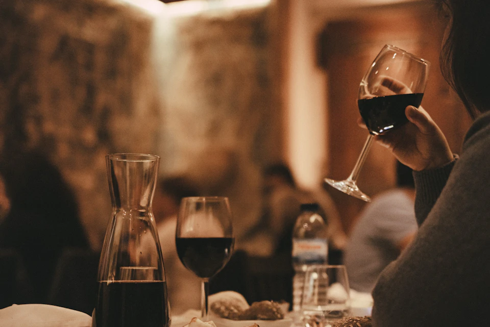 Wine glasses, a carafe of red wine, and a water bottle on a dining table; a hand holding a wine glass in the background.