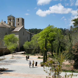 En historisk stenbyggnad med välvda fönster, omgiven av träd och en stenlagd innergård där några personer går.