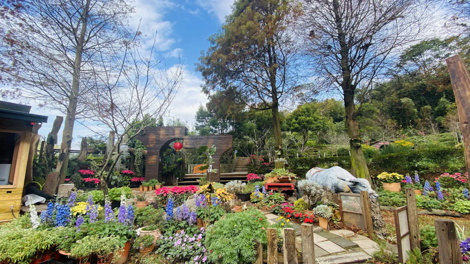 A garden with colorful flowers, trees, a wooden arch, and a stone path under a partly cloudy sky.