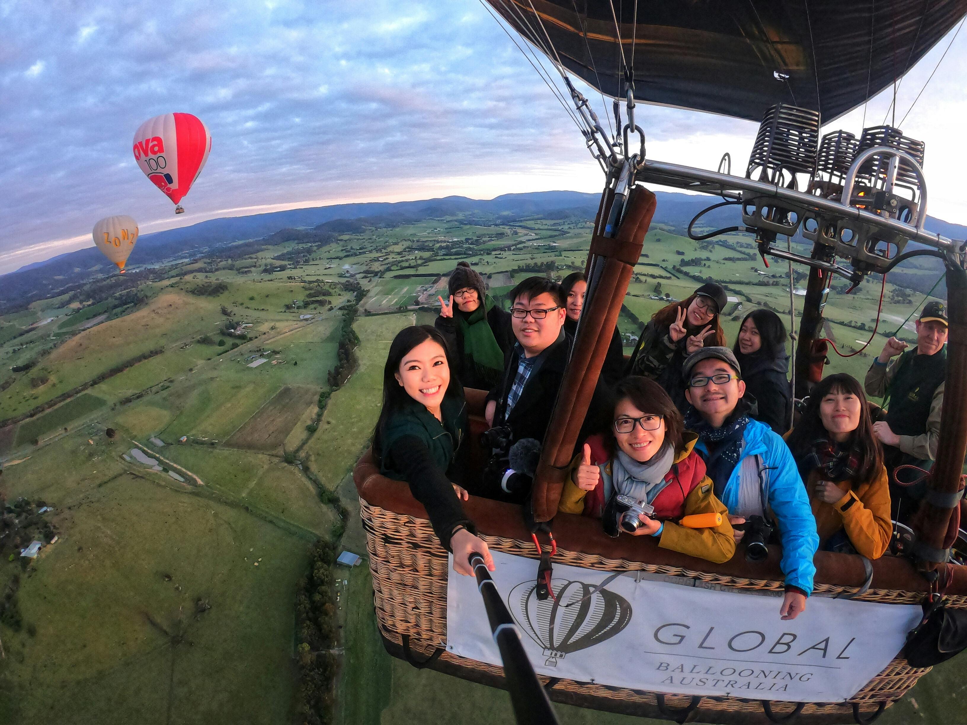 A group of smiling people in a hot air balloon basket with "Global Ballooning Australia" banner, high above a scenic landscape.