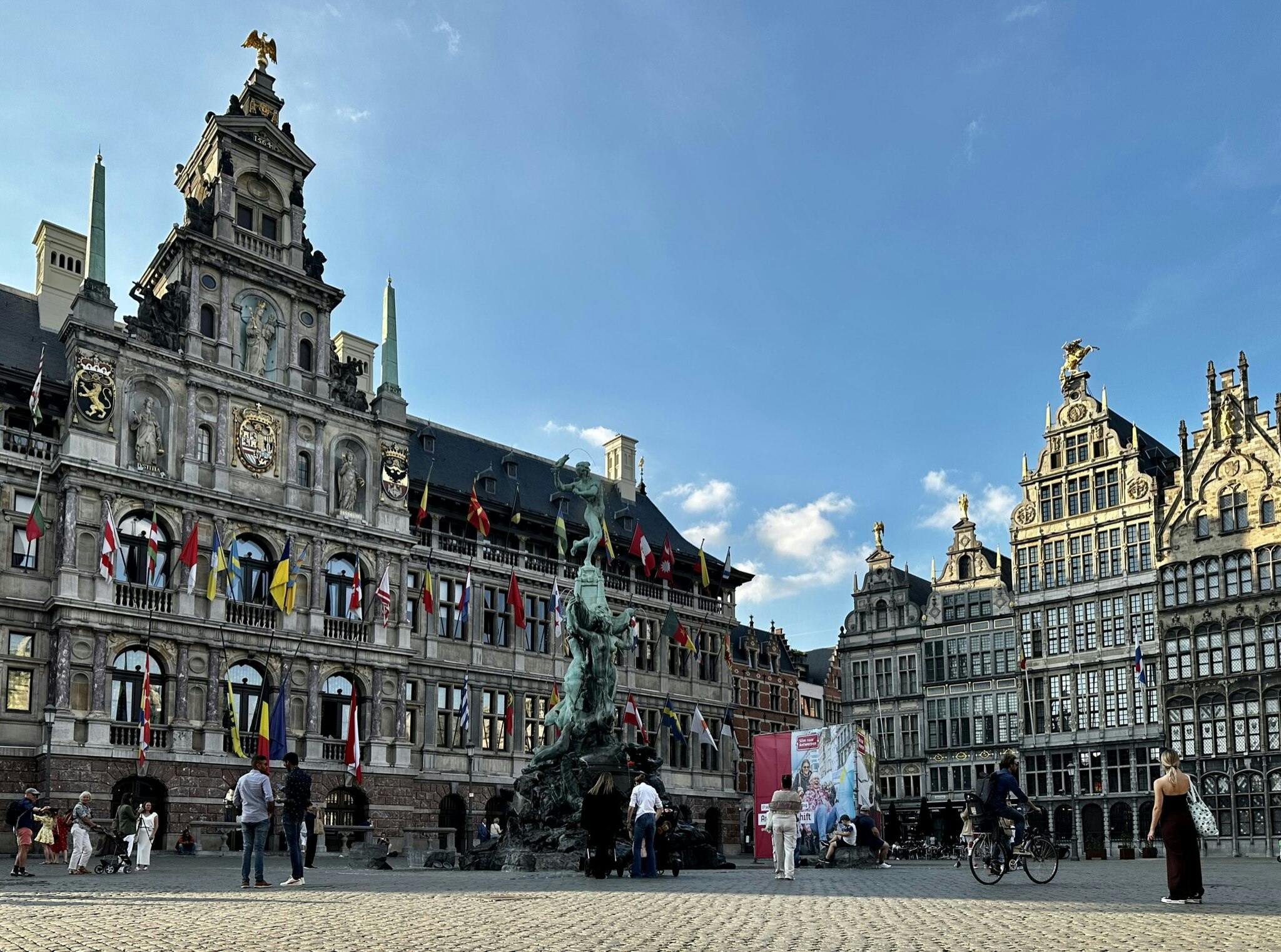 Historické evropské náměstí s ozdobnými budovami, četnými vlajkami, velkou fontánou a lidmi, kteří chodí nebo jezdí na kole.