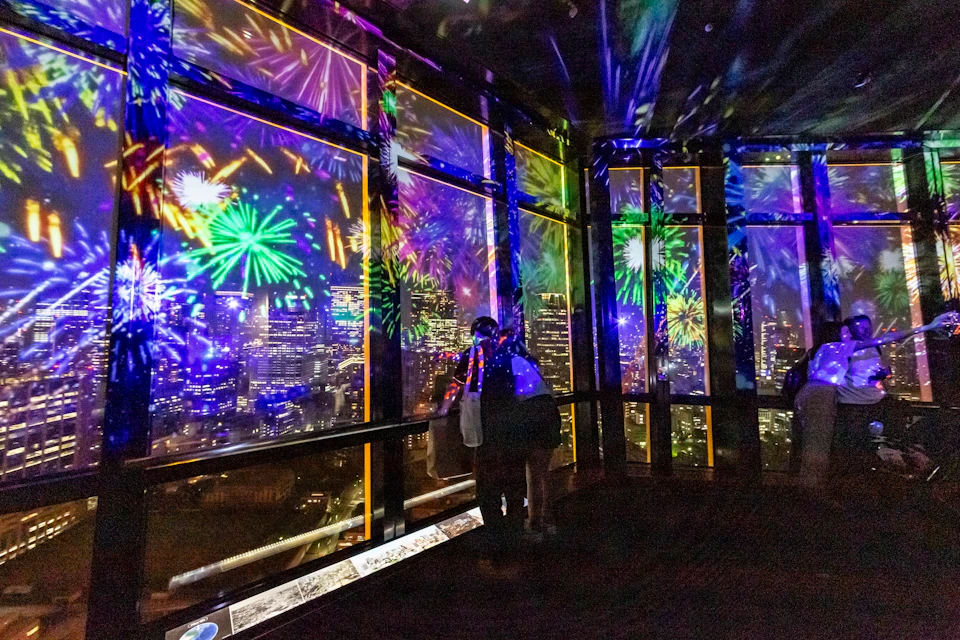 A couple stands in a room with large windows, observing bright fireworks projected over a cityscape at night.