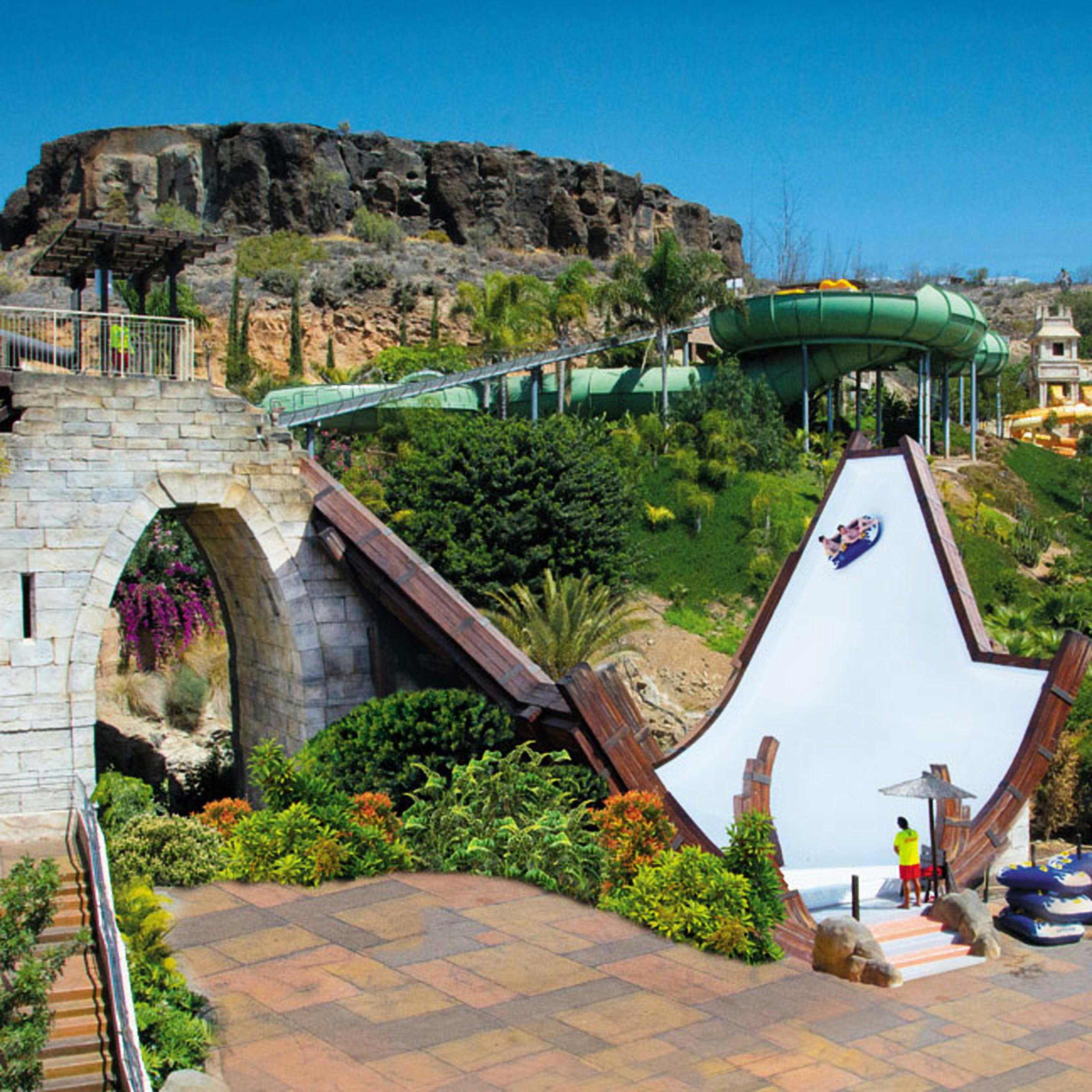 A water park slide propelling a rider vertically, surrounded by lush vegetation and rocky terrain under a clear blue sky.