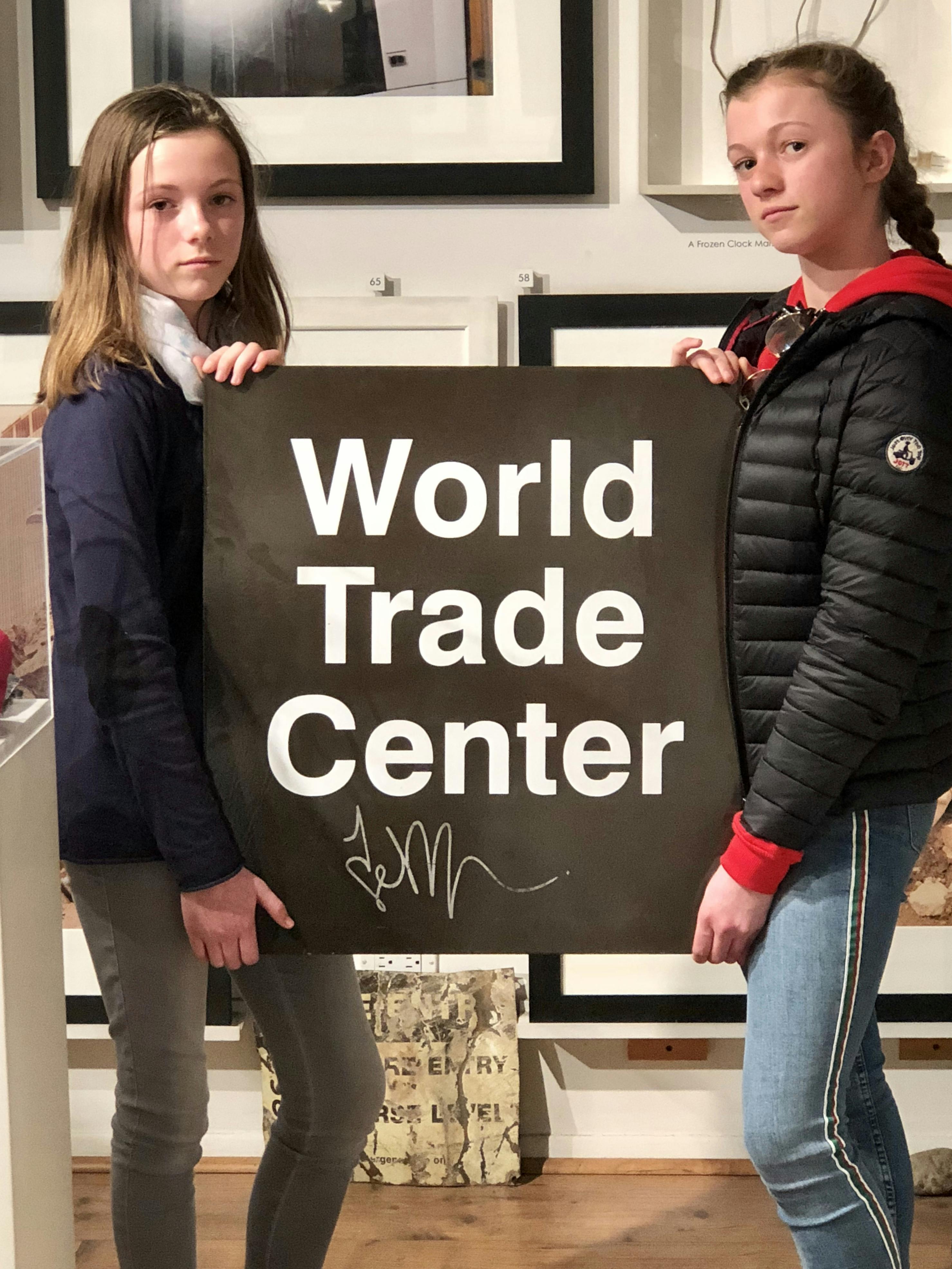 Two girls holding a sign that reads "World Trade Center" with a signature below, standing in a room with framed pictures on the wall.