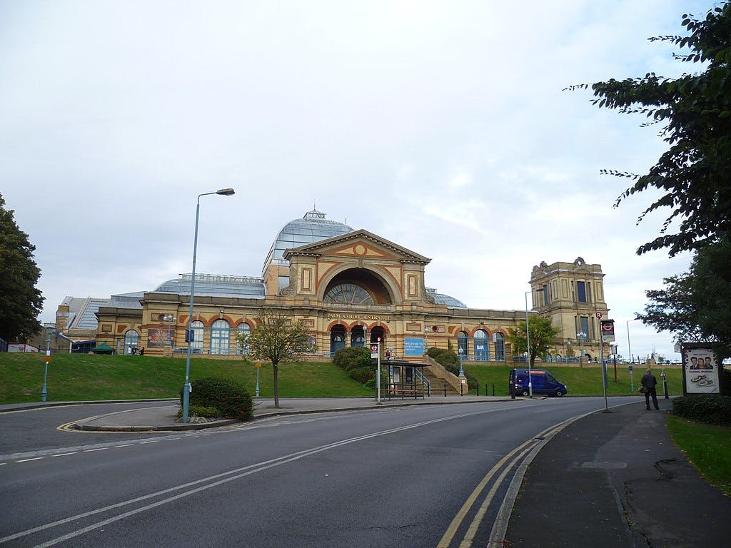 Ein großes historisches Gebäude mit bogenförmigem Eingang, flankiert von Türmen, von der Straße aus gesehen mit einem Lieferwagen und ein paar Fußgängern.