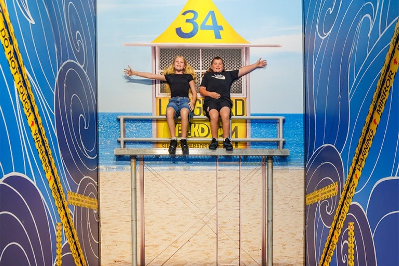 Two children seated on a lifeguard tower set, surrounded by beach-themed decor and mirror illusions.