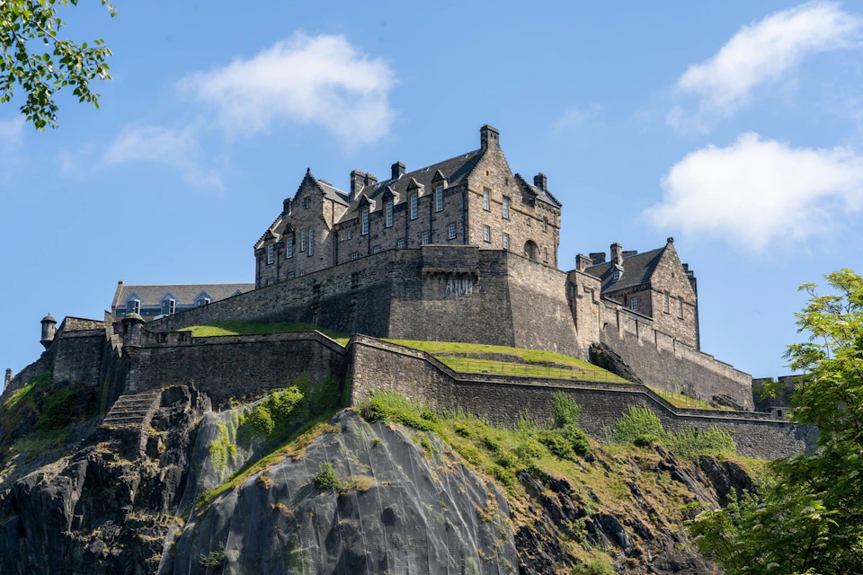 A historic stone castle on a rocky hilltop under a blue sky with scattered clouds.