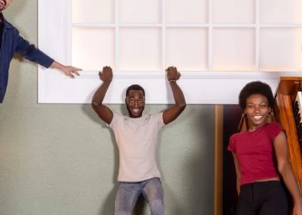 A group of people posing in a room with a tilted perspective, featuring a wall with speakers and a piano on the floor.